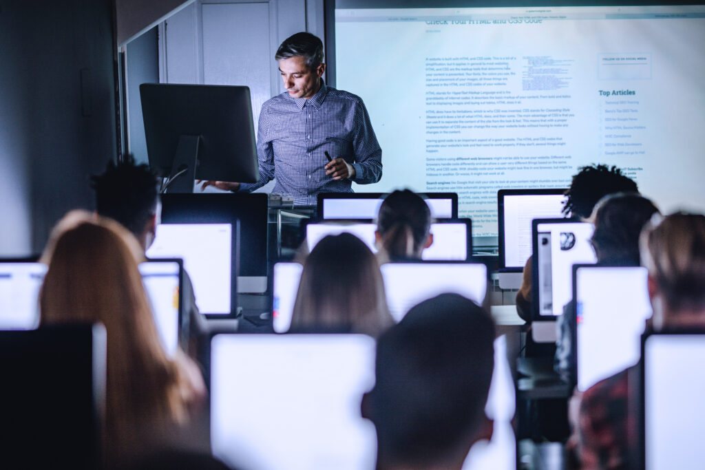Mid Adult Professor Teaching A Lecture From Desktop Pc At Computer Lab.