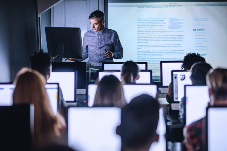 Mid Adult Professor Teaching A Lecture From Desktop Pc At Computer Lab.