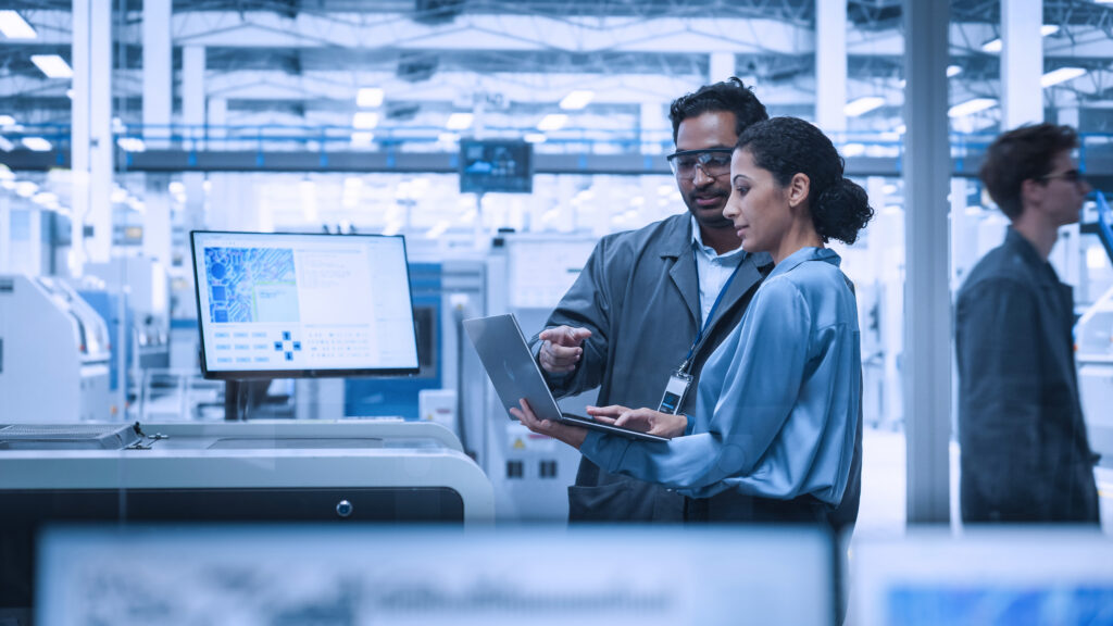 Photograph showing two factory workers discussing data on a laptop in a modern industrial setting with machinery and digital screens in the background.