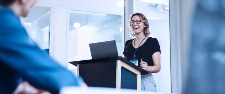 Photograph showing a woman standing behind a podium with a laptop, giving a presentation in a modern office setting.