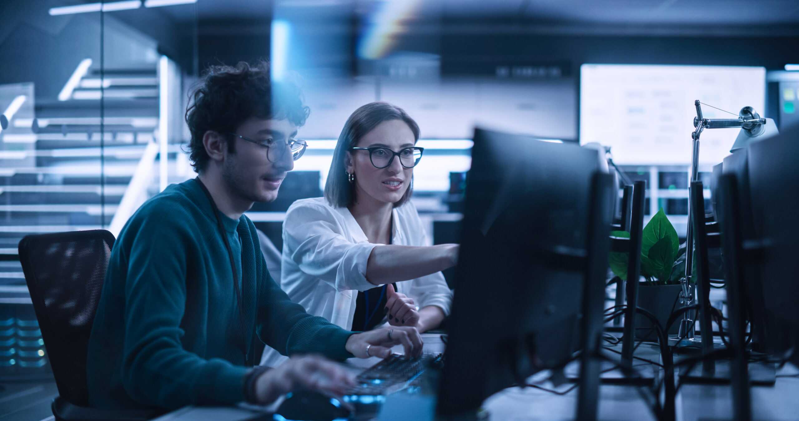 Two Young Colleagues Working On Computers And Talking At A Workp