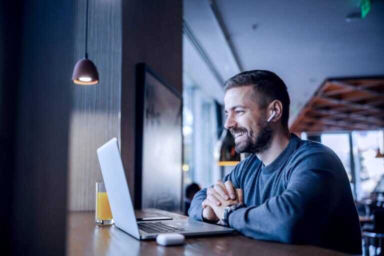 Photograph of a man sitting at a wooden counter in a hotel, working on a laptop with wireless earbuds in his ears.