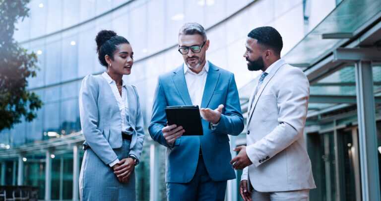 Photograph of three business professionals standing outdoors in front of a modern glass building, engaged in discussion while one person holds a tablet.