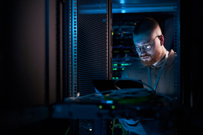 Man working inside a server room, surrounded by network equipment and cables.