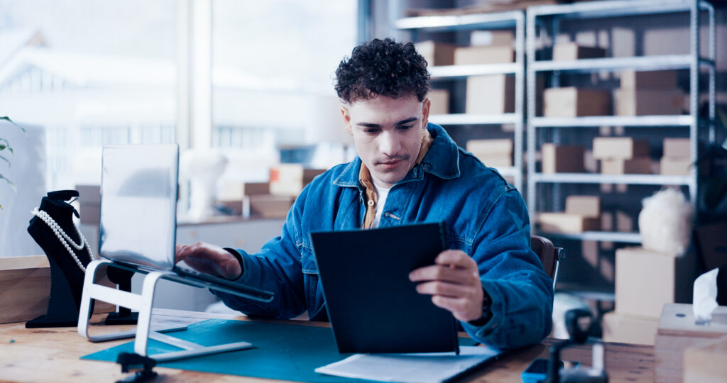 Photograph of a person sitting at a desk in a shop, holding a tablet and using a computer with a monitor on a stand.