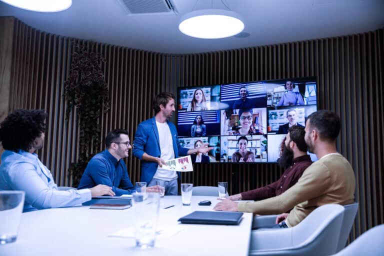 Business meeting with five people seated around a conference table and one person standing, presenting to a large screen displaying a video call with multiple participants.