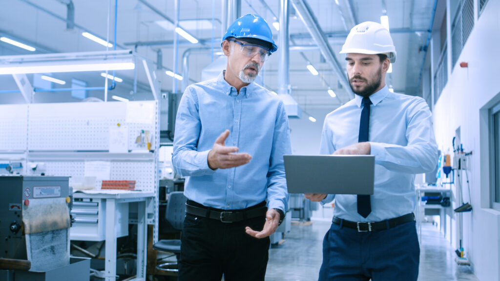 Photograph showing two engineers in a factory setting, both wearing safety helmets and discussing work while one holds a laptop.