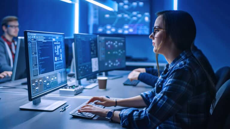 Photograph showing a person working on multiple computer monitors displaying code and data visualisations.