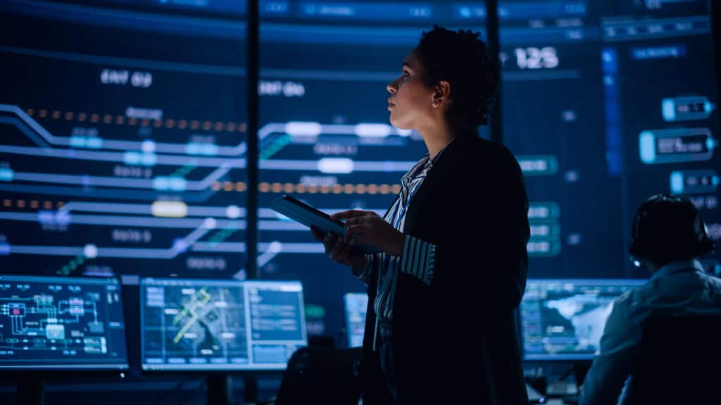 Woman holding a tablet in front of multiple large digital screens displaying complex data visualisations and charts.