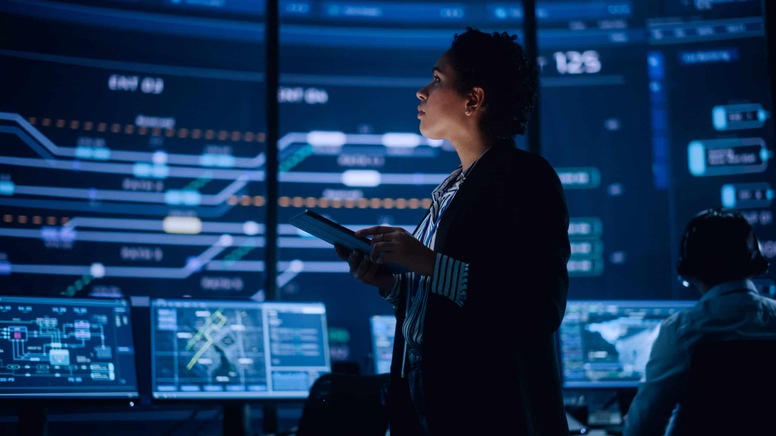 Woman holding a tablet in front of multiple large digital screens displaying complex data visualisations and charts.