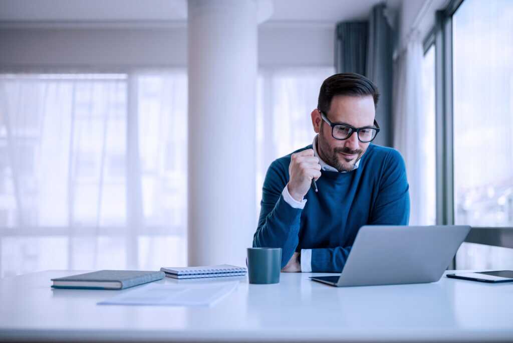 Photograph of a person sitting at a desk with a laptop, notebook, and coffee mug in a bright, modern office space.