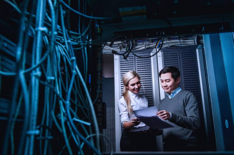 Photograph showing two IT professionals reviewing documents inside a server room surrounded by racks of network equipment and cables.