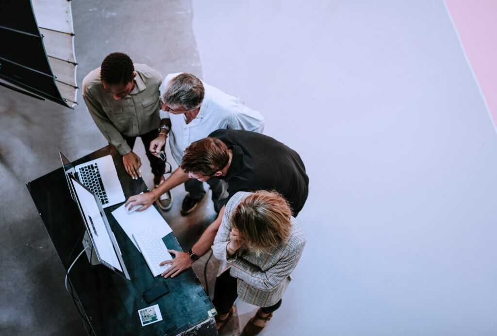 Overhead view of four people gathered around a desk with a laptop and a desktop computer, collaborating on a task.
