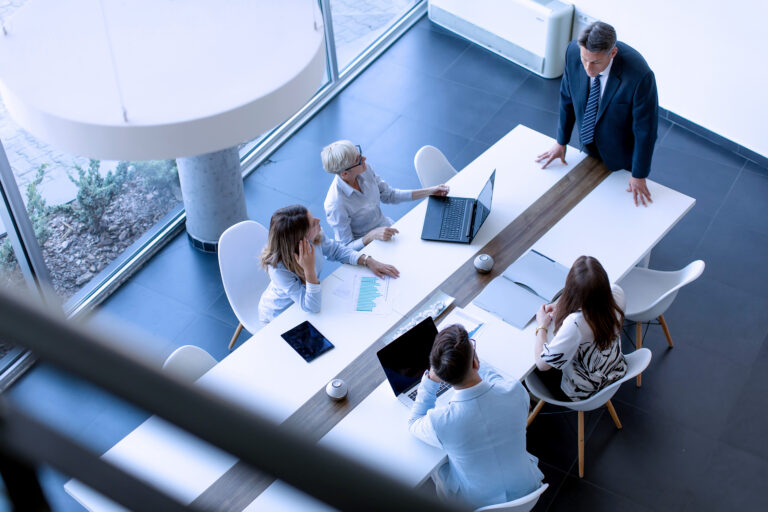 Photograph of a business meeting with five professionals seated around a long white table in a modern office space.