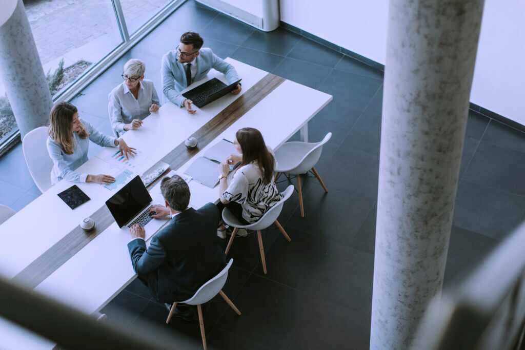 Photograph of a business meeting with five professionals seated around a long white table in a modern office space.