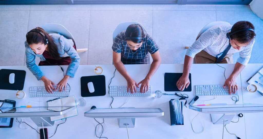 Three people working at a shared desk with desktop computers and keyboards, viewed from above.