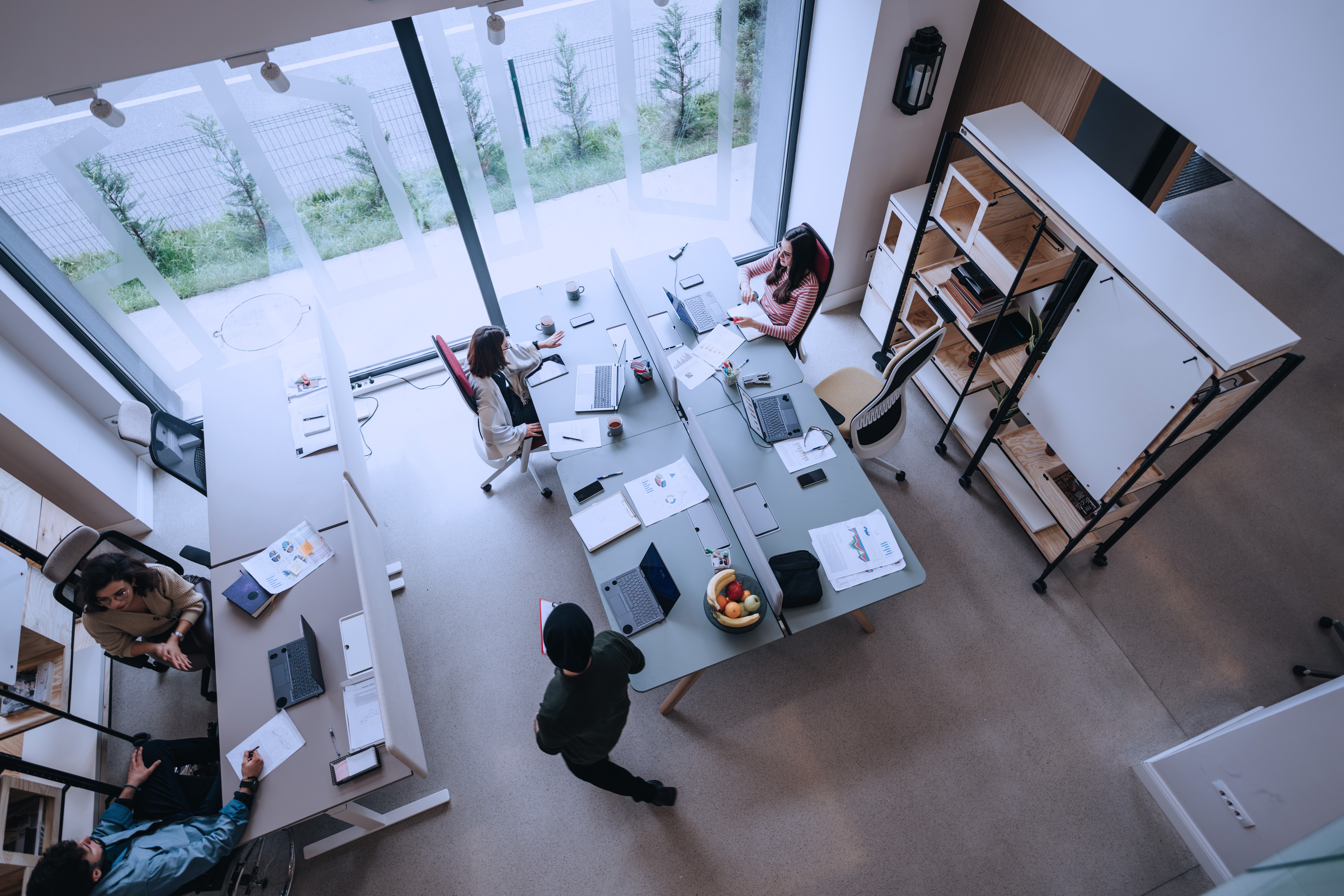 Photograph showing a modern office space with four people engaged in work around two desks arranged in an L-shape near large windows.
