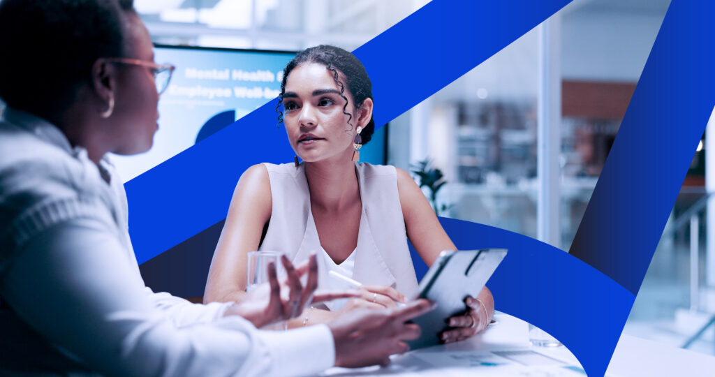 Woman holding a tablet deep in conversation with another woman with the SCC sail graphic in the background.
