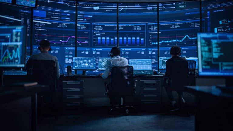 Photograph showing three people seated in a darkened control room facing multiple large screens displaying various financial charts, graphs, and data analytics.