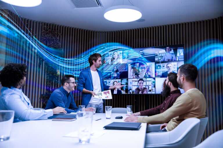 Business meeting with five people seated around a conference table and one person standing, presenting to a large screen displaying a video call with multiple participants. The SCC wave graphic flows in front of the rack.