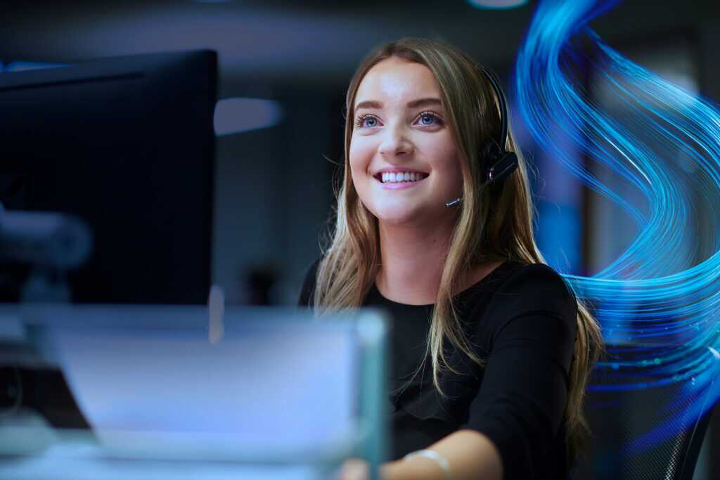 Photograph of a woman wearing a headset and working at a computer in an office setting with the SCC wave graphic in the background.